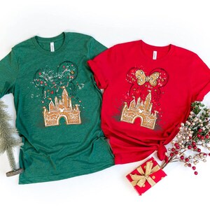 K&ouml;nnte beinhalten: Zwei kurz&auml;rmelige T-Shirts, eines gr&uuml;n und eines rot, mit einem Lebkuchenschloss-Design, Mickey Mouse Ohren und einer Schleife. Die Shirts sind mit festlichen Elementen dekoriert, die ein Feiertags-Thema suggerieren.