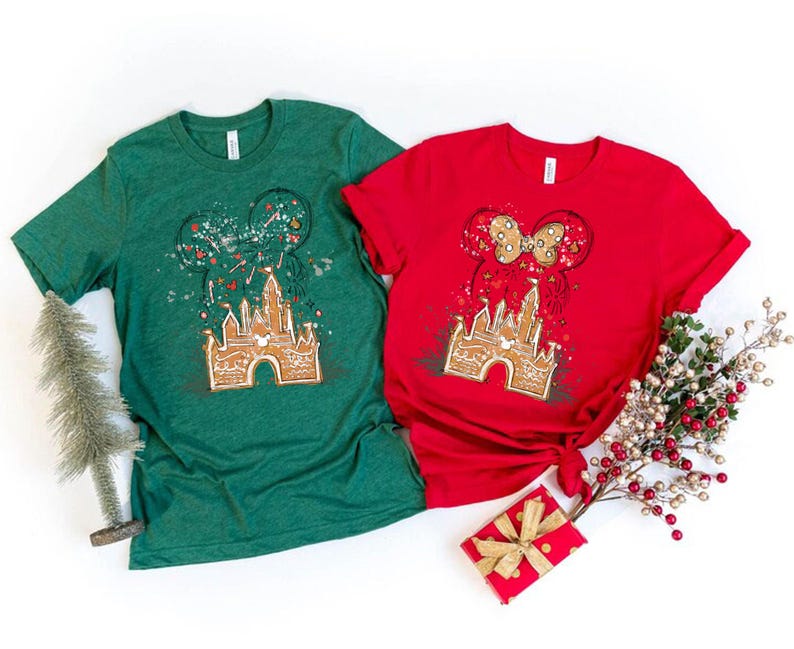 K&ouml;nnte beinhalten: Zwei kurz&auml;rmelige T-Shirts, eines gr&uuml;n und eines rot, mit einem Lebkuchenschloss-Design, Mickey Mouse Ohren und einer Schleife. Die Shirts sind mit festlichen Elementen dekoriert, die ein Feiertags-Thema suggerieren.