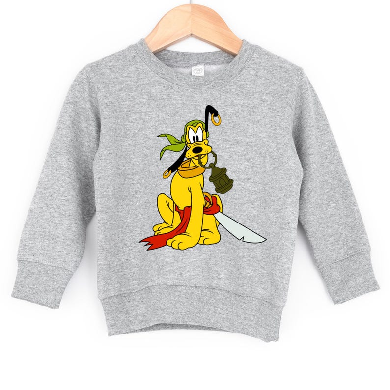 May include: A gray sweatshirt featuring a cartoon Pluto character dressed as a pirate. Pluto is yellow with a green bandana, red sash, and a sword. He holds a lantern and a pirate hook. The sweatshirt is hanging on a wooden hanger.