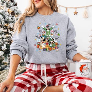 K&ouml;nnte beinhalten: Ein hellgraues Sweatshirt mit einem farbenfrohen Design von Disney-Figuren zum Thema Weihnachten. Das Design zeigt Micky Maus, Minnie Maus, Donald Duck, Daisy Duck und Goofy. Das Sweatshirt wird mit einer rot-wei&szlig; karierten Pyjamahose kombiniert.