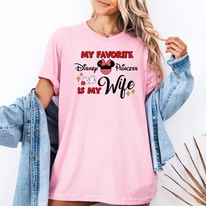 May include: A light pink t-shirt with the text "My Favorite Disney Princess Is My Wife" in red and black. The design includes a Minnie Mouse bow and other graphics. The shirt is short-sleeved.