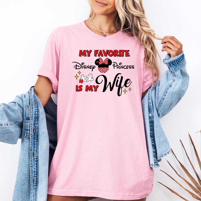 May include: A light pink t-shirt with the text "My Favorite Disney Princess Is My Wife" in red and black. The design includes a Minnie Mouse bow and other graphics. The shirt is short-sleeved.