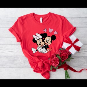 May include: Red t-shirt with a graphic of Mickey Mouse and Minnie Mouse hugging. There are three pink hearts above them.