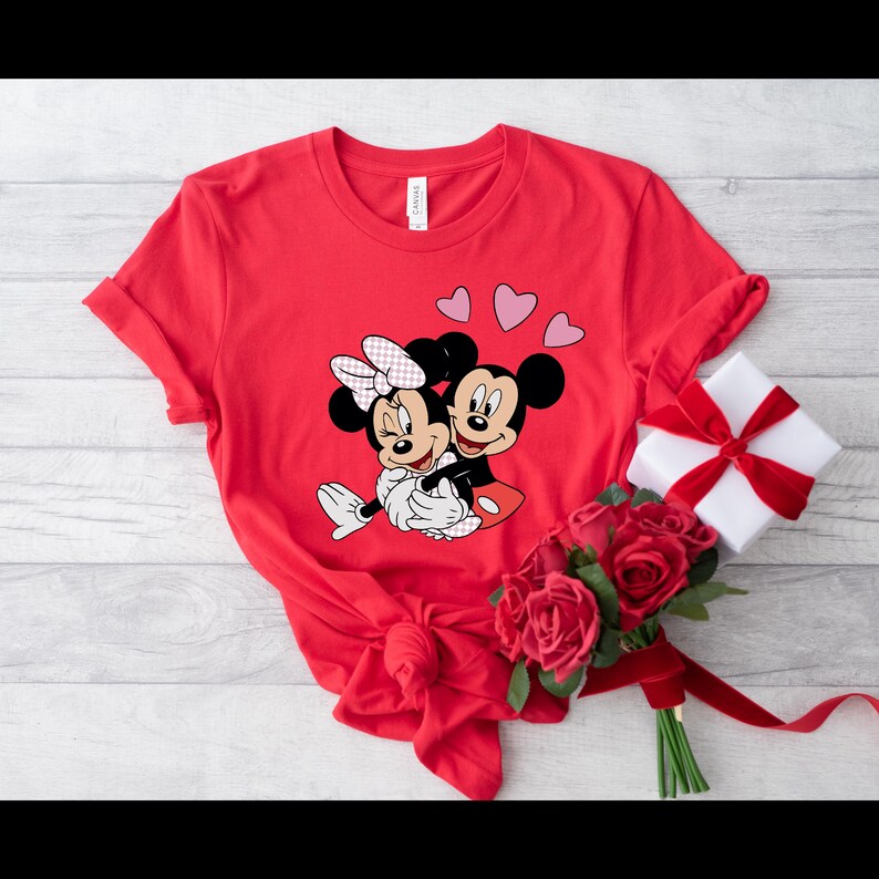 May include: Red t-shirt with a graphic of Mickey Mouse and Minnie Mouse hugging. There are three pink hearts above them.