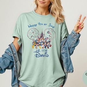 May include: A light green t-shirt with a graphic featuring Mickey Mouse and friends celebrating the 4th of July. The design includes fireworks, a castle, and the text "Happy 4th of July" and "Est. 1776 David".