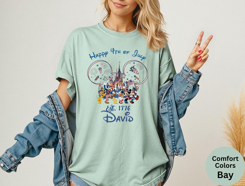 May include: A light green t-shirt with a graphic featuring Mickey Mouse and friends celebrating the 4th of July. The design includes fireworks, a castle, and the text "Happy 4th of July" and "Est. 1776 David".