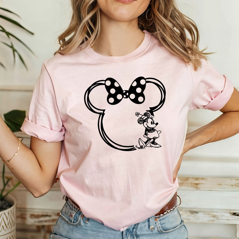 May include: A pink t-shirt with a black and white outline of Mickey Mouse's head and ears. Inside the outline is a black and white image of Minnie Mouse wearing a bow and a dress.