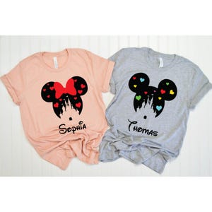 May include: Two light-coloured t-shirts with a black silhouette of Mickey Mouse ears with a castle inside. The left shirt is pink and says "Sophia" and the right shirt is grey and says "Thomas".