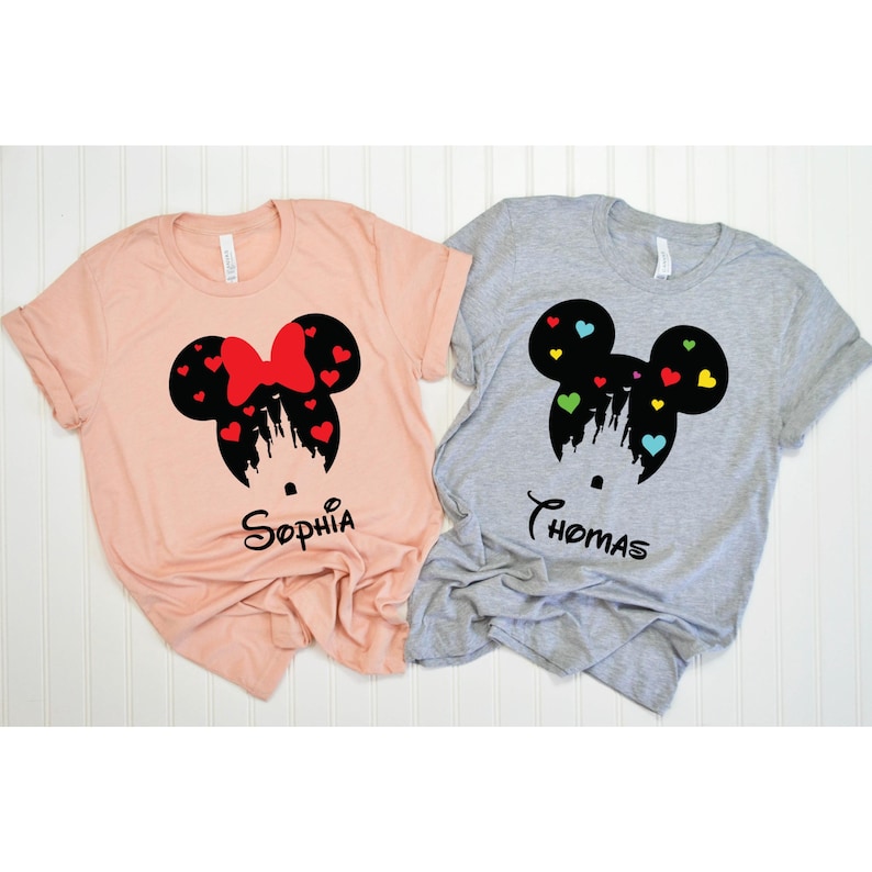 May include: Two light-coloured t-shirts with a black silhouette of Mickey Mouse ears with a castle inside. The left shirt is pink and says "Sophia" and the right shirt is grey and says "Thomas".