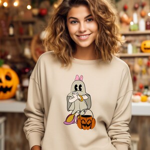 Puede incluir: Una sudadera beige con un fantasma de dibujos animados que lleva un lazo rosa y sostiene un cubo de caramelos. El fantasma lleva una pulsera de oro y tiene una expresi&oacute;n de sorpresa. El fantasma est&aacute; de pie junto a una calabaza con una cara sonriente.