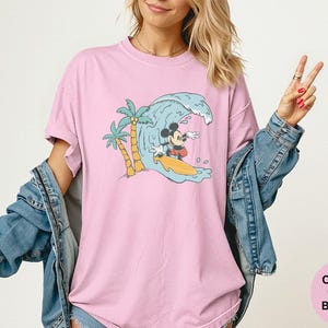 May include: Pink t-shirt with a graphic of Mickey Mouse surfing a wave. The design includes palm trees and a yellow surfboard. The shirt is a Comfort Colors Blossom.