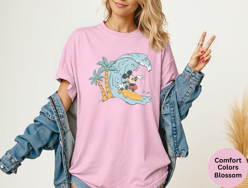 May include: Pink t-shirt with a graphic of Mickey Mouse surfing a wave. The design includes palm trees and a yellow surfboard. The shirt is a Comfort Colors Blossom.