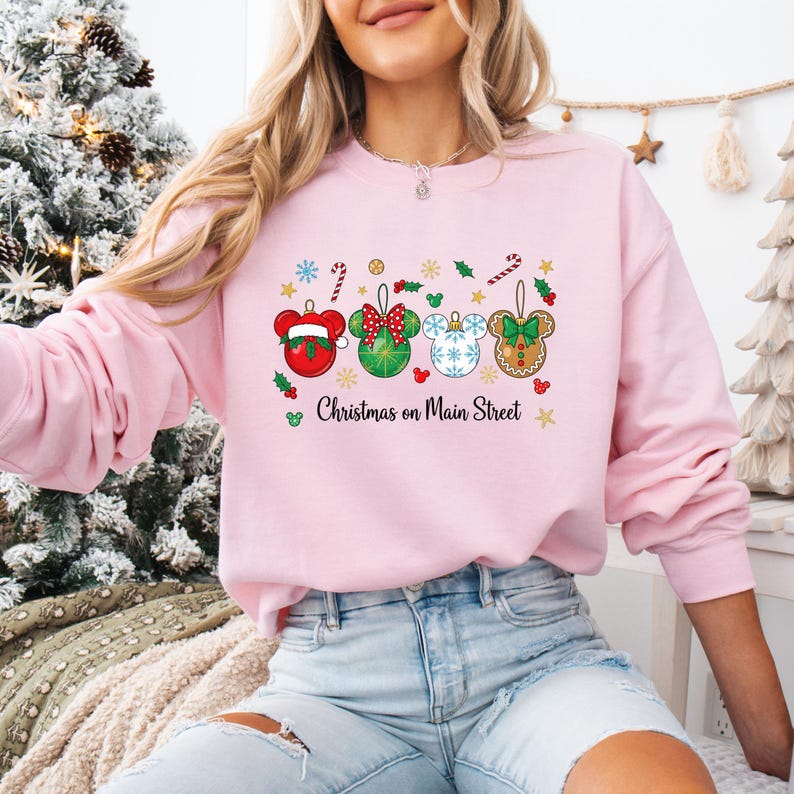 May include: A pale pink sweatshirt with a Christmas-themed design. The design features four Mickey Mouse-shaped baubles in red, green, white, and gingerbread colours, with the text "Christmas on Main Street".