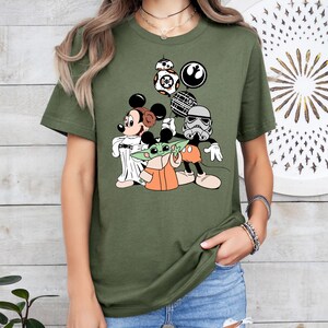 May include: Olive green t-shirt featuring a cartoon graphic of Mickey Mouse, Minnie Mouse, Baby Yoda, and a Stormtrooper. The design includes Star Wars and Disney-themed elements, such as BB-8 and the Rebel Alliance symbol. The characters are in a playful, cartoon style.