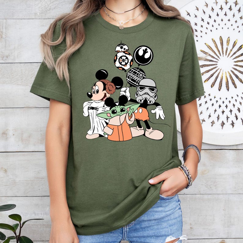 May include: Olive green t-shirt featuring a cartoon graphic of Mickey Mouse, Minnie Mouse, Baby Yoda, and a Stormtrooper. The design includes Star Wars and Disney-themed elements, such as BB-8 and the Rebel Alliance symbol. The characters are in a playful, cartoon style.