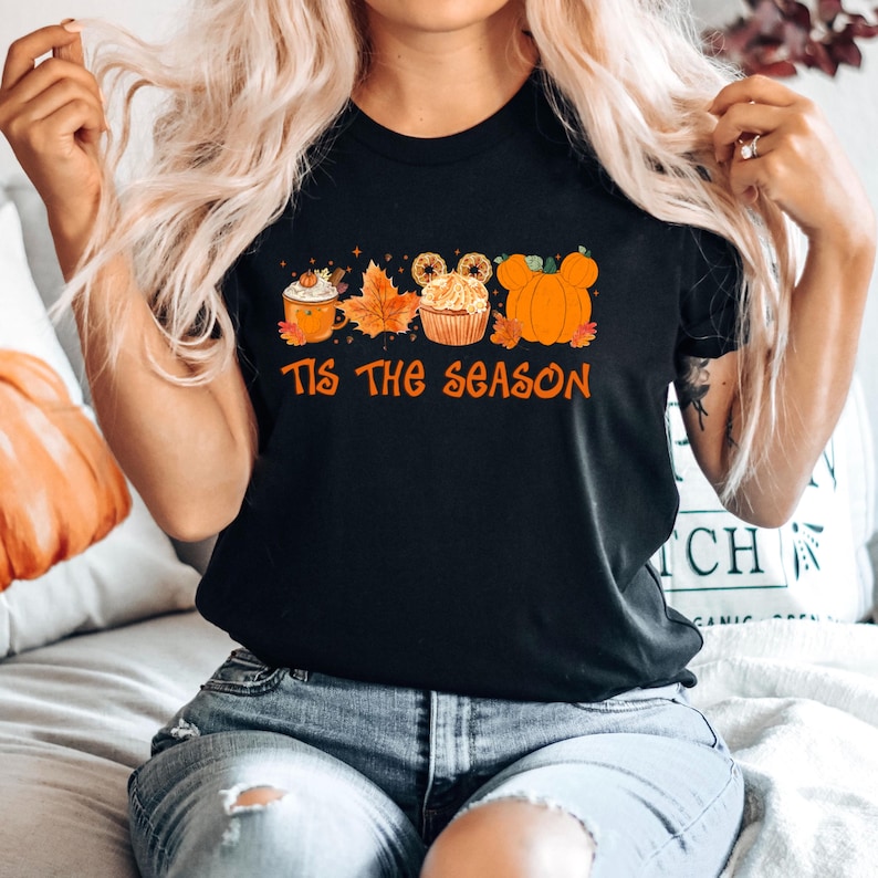 Pu&ograve; includere: T-shirt nero con una grafica arancione che dice "Tis the Season". La grafica include un latte speziato alla zucca, foglie d'autunno, un cupcake con le orecchie di Topolino e una zucca. La maglietta &egrave; indossata da una persona.