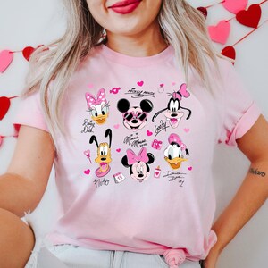 May include: A light pink t-shirt featuring cartoon characters Mickey Mouse, Minnie Mouse, Donald Duck, Daisy Duck, Goofy, and Pluto. The design includes pink hearts and text, perfect for Disney fans.