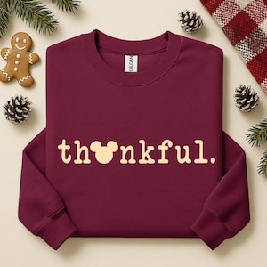 Puede incluir: Una sudadera burdeos con la palabra "thankful" en crema. La "o" de thankful se reemplaza con una cabeza de Mickey Mouse. La sudadera est&aacute; rodeada de pi&ntilde;as, vegetaci&oacute;n y un hombre de jengibre.