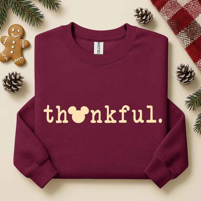 Puede incluir: Una sudadera burdeos con la palabra "thankful" en crema. La "o" de thankful se reemplaza con una cabeza de Mickey Mouse. La sudadera est&aacute; rodeada de pi&ntilde;as, vegetaci&oacute;n y un hombre de jengibre.