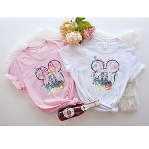 Op de afbeelding: Twee T-shirts met een grafische print van een cartoonkasteel met Mickey Mouse-oren. Het kasteel is roze, blauw en geel. De T-shirts zijn roze en wit.