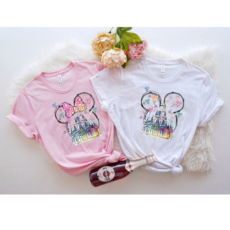 Op de afbeelding: Twee T-shirts met een grafische print van een cartoonkasteel met Mickey Mouse-oren. Het kasteel is roze, blauw en geel. De T-shirts zijn roze en wit.