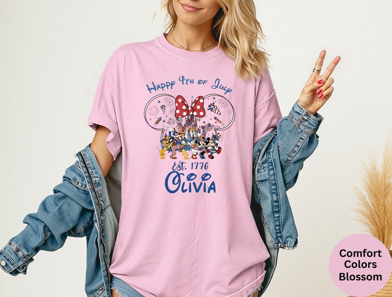 Pode incluir: Camiseta rosa claro com um desenho que inclui orelhas do Mickey, um la&ccedil;o vermelho e o texto "Happy 4th of July Est. 1776 Olivia". O design inclui personagens de desenhos animados e fogos de artif&iacute;cio.