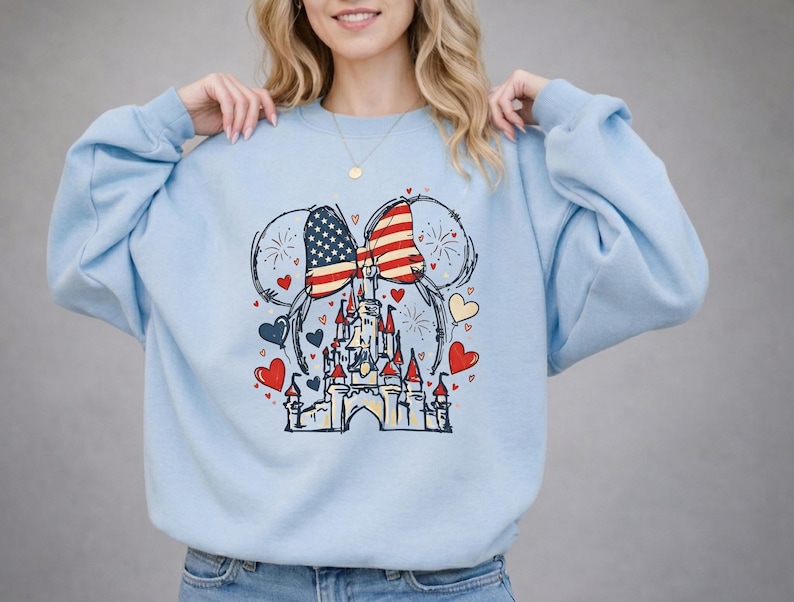 May include: Light blue sweatshirt featuring a patriotic design. The graphic includes a bow with the American flag, fireworks, hearts, and a castle. The design is in red, white, blue, and black.
