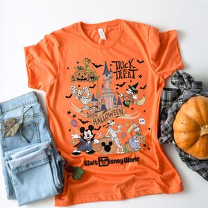 May include: Orange t-shirt with a Mickey Mouse Halloween design. The shirt features Mickey Mouse, Goofy, and other Disney characters dressed in Halloween costumes. The text "Trick or Treat" and "2024 Halloween" are printed on the shirt. The shirt also features a graphic of Cinderella's castle and a pumpkin. The text "Walt Disney World" is printed at the bottom of the shirt.