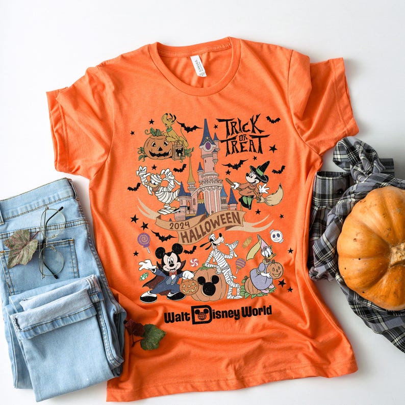 May include: Orange t-shirt with a Mickey Mouse Halloween design. The shirt features Mickey Mouse, Goofy, and other Disney characters dressed in Halloween costumes. The text "Trick or Treat" and "2024 Halloween" are printed on the shirt. The shirt also features a graphic of Cinderella's castle and a pumpkin. The text "Walt Disney World" is printed at the bottom of the shirt.