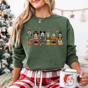 May include: Green sweatshirt featuring a colorful graphic of Disney characters as nutcrackers. The characters include Mickey Mouse, Minnie Mouse, Donald Duck, and Goofy, each in festive outfits. The design is set against a green background, perfect for the holiday season.