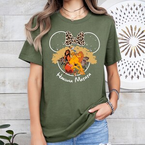 Puede incluir: Camiseta verde con un contorno blanco de las orejas de Mickey Mouse y un lazo de estampado de leopardo. Dentro de las orejas hay una escena de El Rey Le&oacute;n con Simba, Nala, Tim&oacute;n y Pumba. El texto "Hakuna Matata" est&aacute; impreso debajo de la imagen.