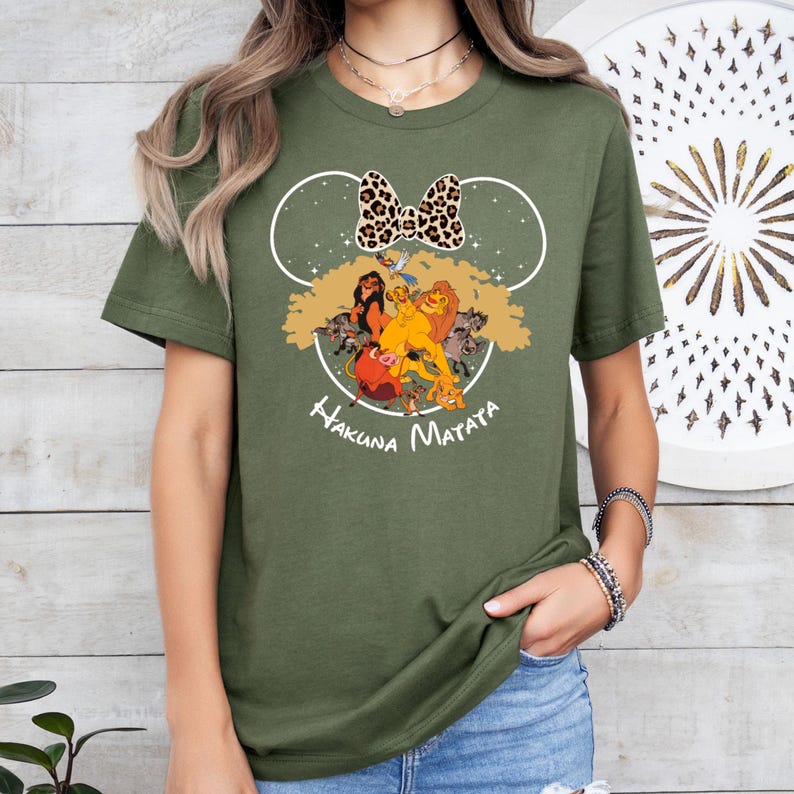Puede incluir: Camiseta verde con un contorno blanco de las orejas de Mickey Mouse y un lazo de estampado de leopardo. Dentro de las orejas hay una escena de El Rey Le&oacute;n con Simba, Nala, Tim&oacute;n y Pumba. El texto "Hakuna Matata" est&aacute; impreso debajo de la imagen.
