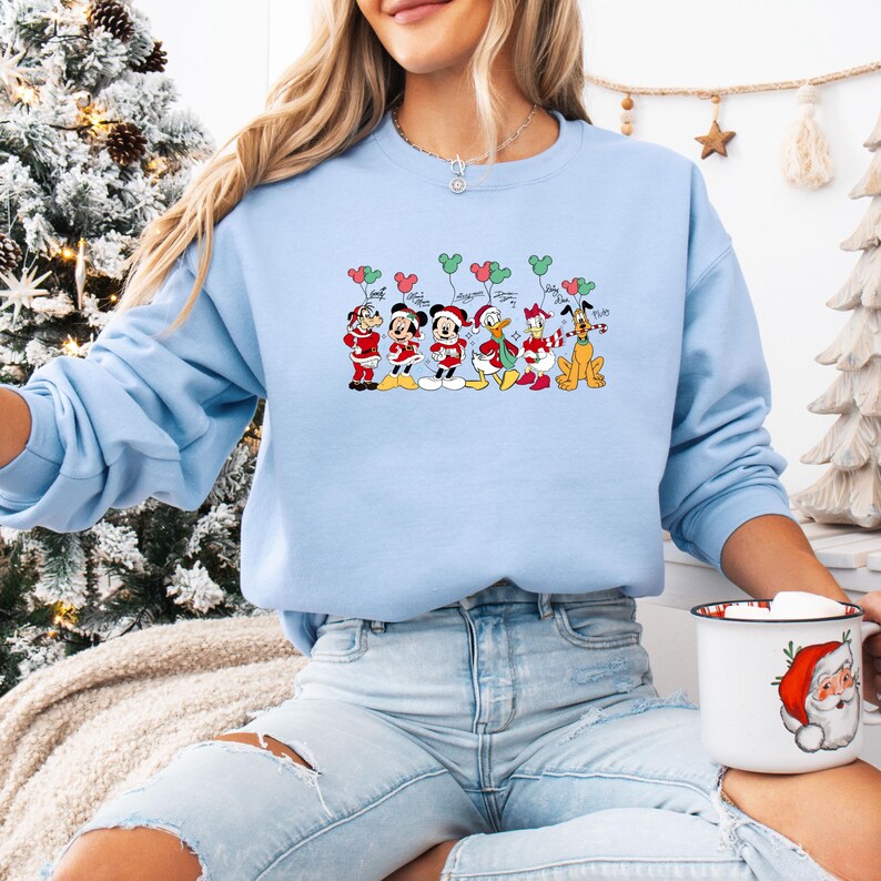 May include: A light blue sweatshirt featuring a festive cartoon design. The graphic depicts Mickey Mouse, Minnie Mouse, Donald Duck, Daisy Duck, and Pluto in Santa Claus outfits, holding balloons. The design is perfect for the holiday season.