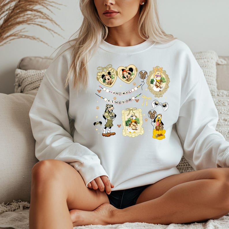May include: White sweatshirt featuring colorful Disney character graphics. The design includes Mickey and Minnie Mouse in heart-shaped frames, framed character portraits, and the text "ANIMAL KINGDOM" on a banner.
