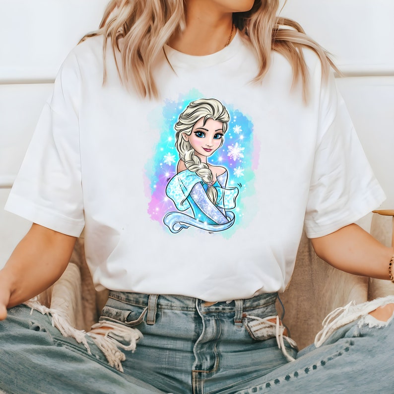 Op de afbeelding: Wit T-shirt met een kleurrijke illustratie van een cartoonprinses met lang blond haar en een blauwe jurk. De prinses is omringd door sneeuwvlokken en een blauw lint.