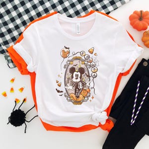 May include: White t-shirt with a Halloween-themed graphic featuring a cartoon mouse dressed as a skeleton. The design includes candy corn, bats, and the word "Spooky". The shirt has orange trim and is paired with black pants.