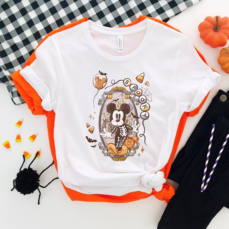 May include: White t-shirt with a Halloween-themed graphic featuring a cartoon mouse dressed as a skeleton. The design includes candy corn, bats, and the word "Spooky". The shirt has orange trim and is paired with black pants.