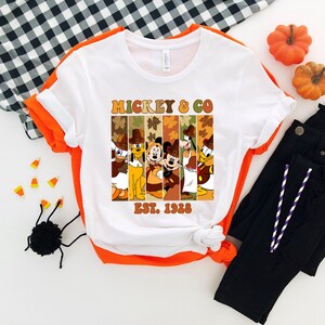 May include: White t-shirt with orange trim featuring a graphic of Mickey & Co characters in fall attire. The design includes the text "Mickey & Co" and "Est. 1928". The shirt is paired with black pants, pumpkins, and Halloween decorations.