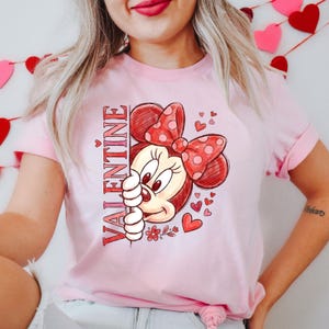 May include: A pale pink t-shirt with a Valentine's Day design. The graphic features a cartoon character with a red bow and the word "VALENTINE" in red. Small red hearts and flowers are also included in the design.