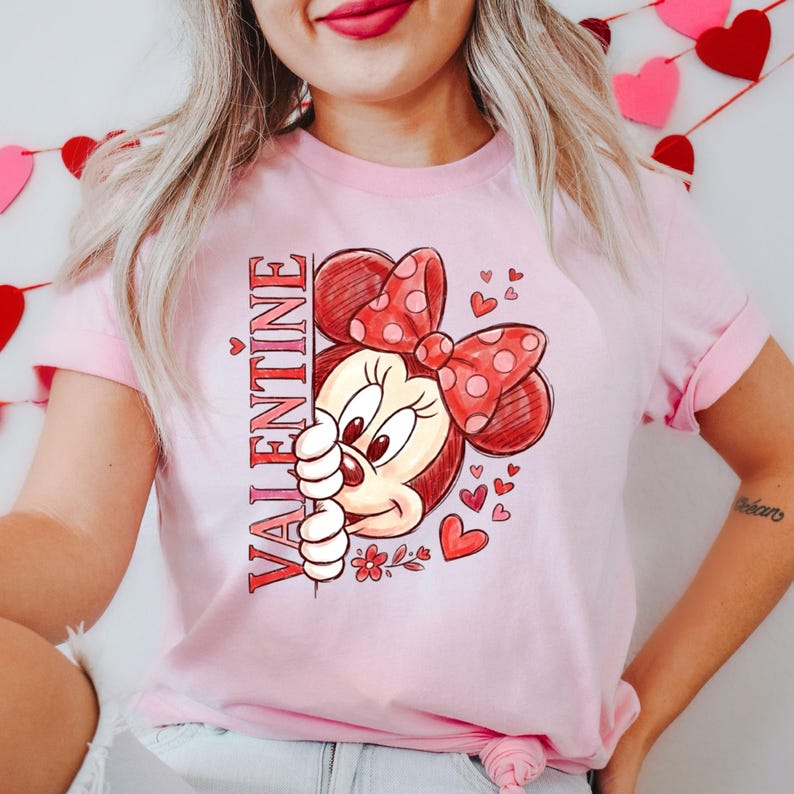 May include: A pale pink t-shirt with a Valentine's Day design. The graphic features a cartoon character with a red bow and the word "VALENTINE" in red. Small red hearts and flowers are also included in the design.