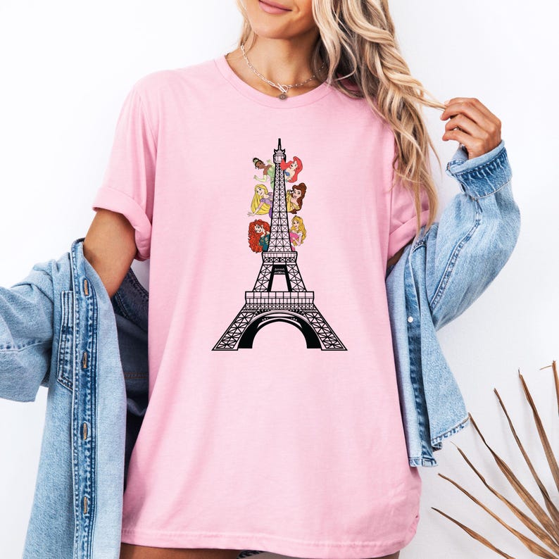 May include: Pink t-shirt featuring a black and white Eiffel Tower design with cartoon princess characters. The shirt has short sleeves and a crew neck. The design is centered on the front of the shirt.