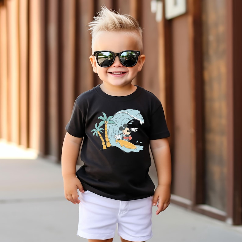May include: A young boy wearing a black t-shirt with a graphic of Mickey Mouse surfing on a wave. The shirt also features palm trees. The boy is wearing white shorts and black sunglasses.