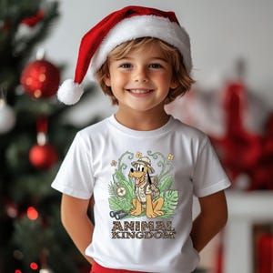 May include: A child wearing a white t-shirt with a cartoon illustration of Pluto in safari attire, the words "ANIMAL KINGDOM" are below the illustration. The child is also wearing a red and white Santa hat. A Christmas tree is in the background.