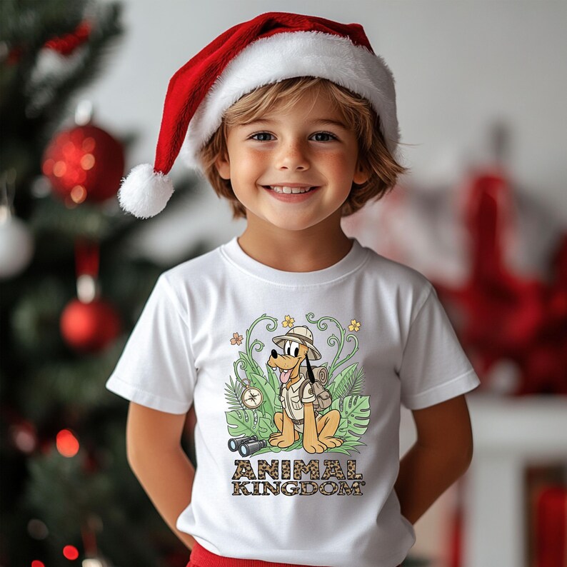 May include: A child wearing a white t-shirt with a cartoon illustration of Pluto in safari attire, the words "ANIMAL KINGDOM" are below the illustration. The child is also wearing a red and white Santa hat. A Christmas tree is in the background.