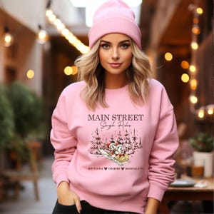 May include: A light pink sweatshirt and beanie. The sweatshirt features the text "Main Street Sleigh Rides" with a graphic of Mickey and Minnie Mouse sledding. The bottom of the graphic reads "Hot Cocoa Cookies Magical Fun."