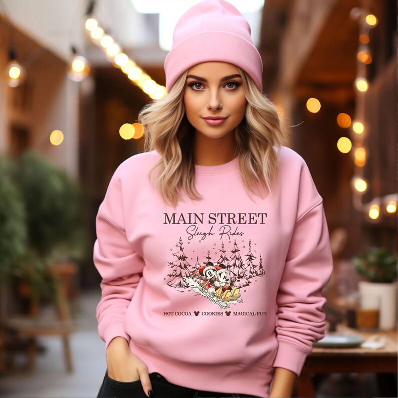 May include: A light pink sweatshirt and beanie. The sweatshirt features the text "Main Street Sleigh Rides" with a graphic of Mickey and Minnie Mouse sledding. The bottom of the graphic reads "Hot Cocoa Cookies Magical Fun."