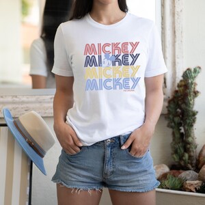 May include: White t-shirt with the word "MICKEY" stacked four times in red, blue, yellow, and light blue. The word "MICKEY" has a retro font. A small Mickey Mouse graphic is in the second "MICKEY". The text "THE ORIGINAL" is below the last "MICKEY".