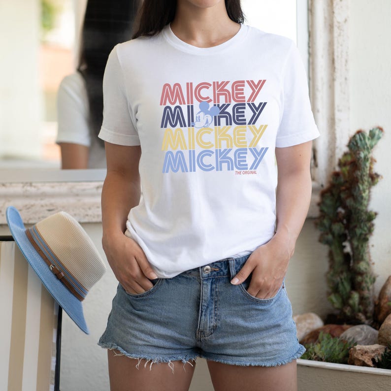 May include: White t-shirt with the word "MICKEY" stacked four times in red, blue, yellow, and light blue. The word "MICKEY" has a retro font. A small Mickey Mouse graphic is in the second "MICKEY". The text "THE ORIGINAL" is below the last "MICKEY".