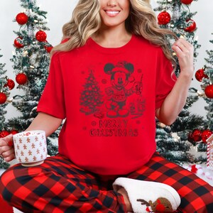 May include: A red t-shirt with a graphic of Mickey Mouse holding a drink, a Christmas tree, and the words "Merry Christmas." The person is wearing red and black plaid pajama pants and white slippers.
