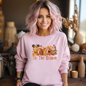 K&ouml;nnte beinhalten: Hellrosa Sweatshirt mit einem herbstlichen Motiv. Die Grafik zeigt ein Getr&auml;nk im Mickey-Mouse-Stil, K&uuml;rbisse, einen Football und eine Tasse Schlagsahne. Der Text "Tis The Season" ist darunter gedruckt.